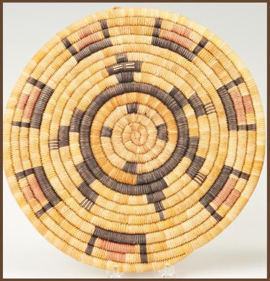 A circular woven basket with geometric patterns, featuring natural and dyed fibers in beige, brown, and black colors.